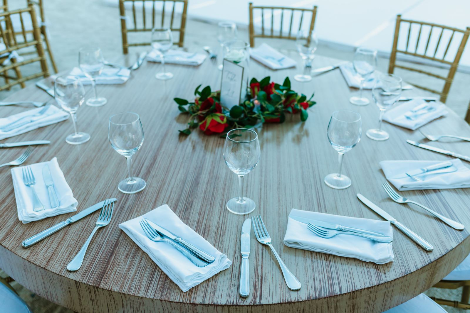 A round reception table set with glassware, folded napkins and a red rose centrepiece
